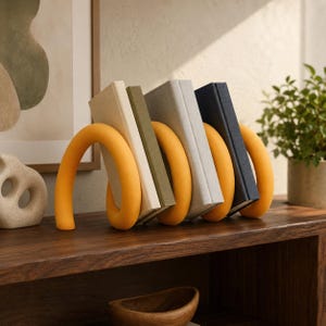 Puede incluir: Una estructura de sujetalibros naranja mate, curva y continua, que sostiene cuatro libros. Los libros tienen cubiertas lisas en tonos crema, verde oliva, gris claro y azul marino oscuro, dispuestos verticalmente dentro del marco de soporte.