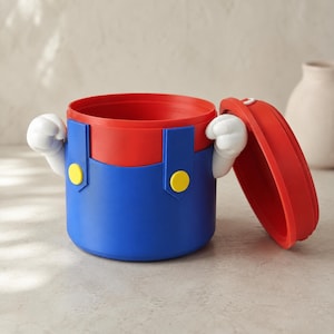 Puede incluir: Un recipiente cilíndrico con una sección superior roja y una sección inferior azul con dos botones circulares amarillos. El recipiente tiene dos manos blancas con guantes de estilo caricatura a los lados y una tapa roja a juego.