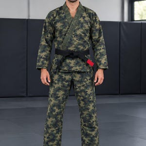 Op de afbeelding: Een jiu-jitsu gi met camouflageprint bestaande uit een jas met lange mouwen en een overslag aan de voorzijde, gecombineerd met een bijpassende broek. De set wordt in de taille vastgezet met een zwarte riem met een klein rood accent.