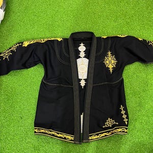 Puede incluir: Una chaqueta de artes marciales negra con intrincados bordados dorados en las mangas, el pecho y la espalda. La prenda tiene un cuello reforzado con costuras doradas y el dobladillo está decorado con un patrón dorado detallado en la parte inferior.