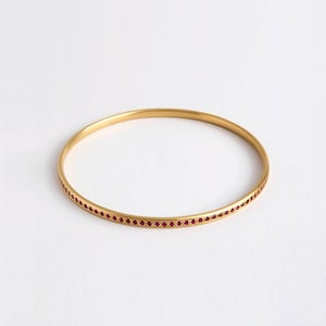 Puede incluir: Un brazalete de oro con una sola fila de pequeñas piedras preciosas rojas alrededor del borde.