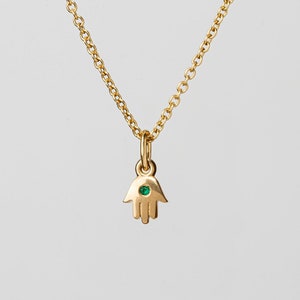 Puede incluir: Un collar de cadena de oro con un pequeño dije de mano de hamsa de oro con una piedra preciosa verde en el centro.