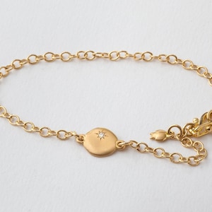 Könnte beinhalten: Ein goldenes Kettenarmband mit einem runden Anhänger, der einen kleinen Diamanten in der Mitte trägt.
