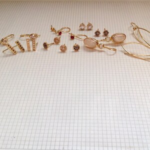 May include: A collection of gold earrings with various styles and gemstones, including a pair with a light pink oval stone, a pair with a small round stone, and a pair with a small black stone.