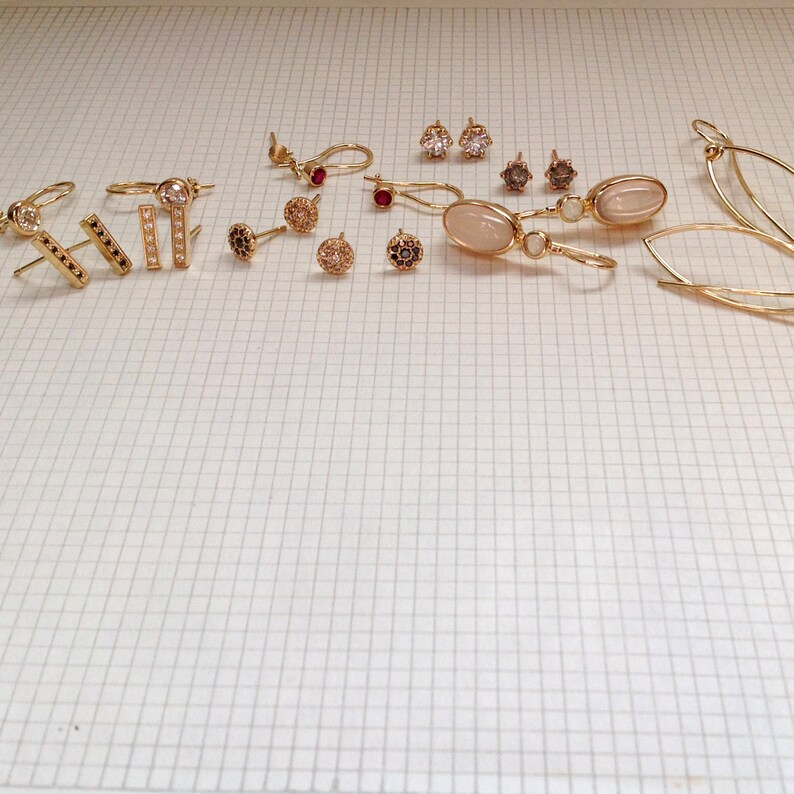 May include: A collection of gold earrings with various styles and gemstones, including a pair with a light pink oval stone, a pair with a small round stone, and a pair with a small black stone.