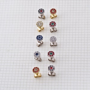May include: Ten pairs of small, round earrings with a flower design. Each earring has a center stone surrounded by smaller stones. The center stones are red, blue, green, or clear. The earrings are on a white background.
