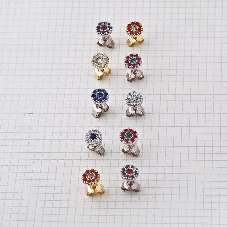 May include: Ten pairs of small, round earrings with a flower design. Each earring has a center stone surrounded by smaller stones. The center stones are red, blue, green, or clear. The earrings are on a white background.