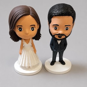 Puede incluir: Dos figuras cabezonas personalizadas sobre bases circulares blancas. Una figura tiene cabello castaño hasta los hombros y viste un vestido blanco sin mangas. La otra tiene cabello negro corto, barba poblada y viste un traje oscuro con camisa blanca.