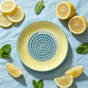 May include: A round ceramic grater plate with a pale yellow rim and a central blue-toned, textured circular pattern for grating. The plate is surrounded by several fresh lemon slices and green mint leaves on a light blue fabric surface.