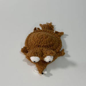 May include: A handmade brown crochet creature with large white eyes and a pointed nose. The whimsical design features a textured body and a small white detail at the nose, creating a unique, playful aesthetic. The creature has a tuft of brown yarn on its head.