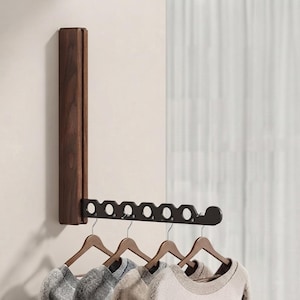 May include: A wall-mounted folding clothes rack featuring a vertical dark brown wooden base and a horizontal black metal arm with six circular cutouts for hangers. Four wooden hangers with garments are suspended from the metal arm.