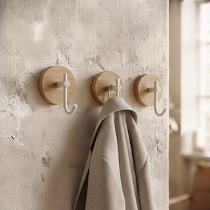 Könnte beinhalten: Drei Wandhaken mit runden Sockeln aus hellem Holz und weißen, J-förmigen Metallhaken. Ein hellbeiger Mantel hängt am mittleren Haken. Die Haken sind an einer strukturierten, hellen Wandfläche in einer vertikalen Anordnung befestigt.