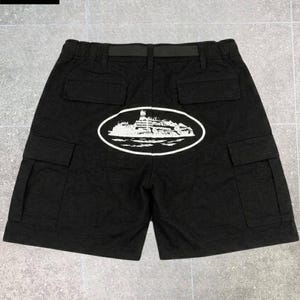 May include: Black cargo shorts featuring an elastic waistband with an integrated black belt. The back displays a white oval graphic containing an illustration of a large ship on water. The shorts include two back pockets and two side cargo pockets.