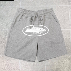 May include: Grey shorts featuring an elastic waistband with a drawstring and a white graphic print of a cruise ship inside an oval frame on the front. The shorts include two side pockets and are made of a soft, heathered fabric material.