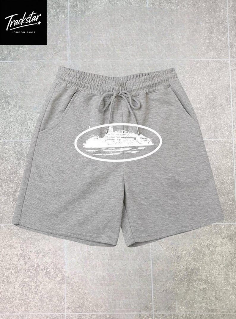 May include: Grey shorts featuring an elastic waistband with a drawstring and a white graphic print of a cruise ship inside an oval frame on the front. The shorts include two side pockets and are made of a soft, heathered fabric material.