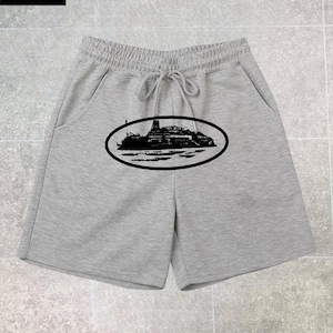 May include: Light gray shorts featuring an elastic waistband with a drawstring and a black oval graphic on the front. The graphic depicts a silhouette of a large ship on the water. The shorts have two side pockets and a straight hemline.