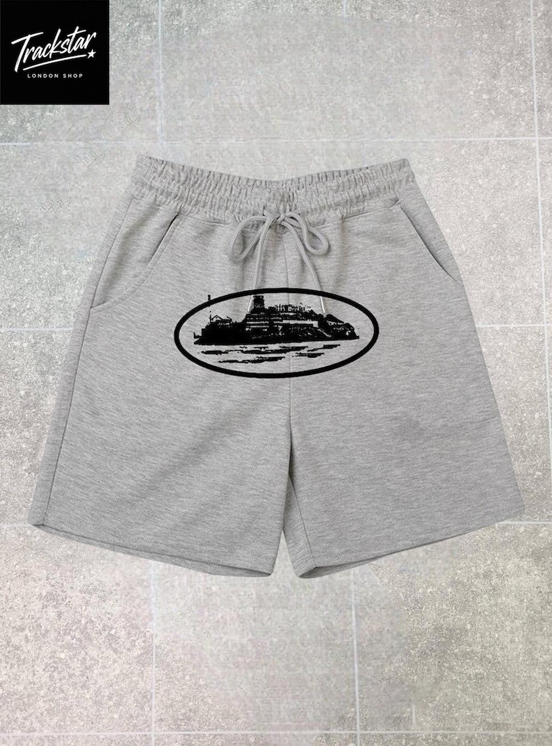 May include: Light gray shorts featuring an elastic waistband with a drawstring and a black oval graphic on the front. The graphic depicts a silhouette of a large ship on the water. The shorts have two side pockets and a straight hemline.