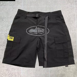 May include: Black cargo shorts featuring an integrated adjustable belt with a buckle, two side cargo pockets, and a yellow rectangular patch with the text LITEWORK. An oval graphic print of a ship is centered on the front of the shorts.