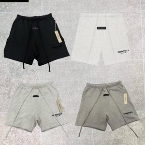 Op de afbeelding: Vier paar sportshorts in zwart, wit en twee grijstinten. Elke broek heeft een trekkoord, een kleine rechthoekige patch en het woord "ESSENTIALS" op de pijp gedrukt. Het logo van Trackstar London Shop staat linksboven.