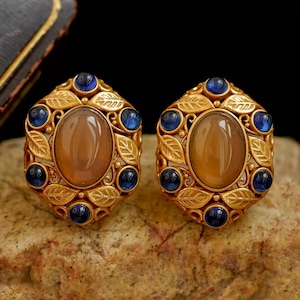 May include: A pair of gold-tone earrings featuring a central oval cabochon in a warm tan hue, surrounded by a decorative frame of embossed leaves and six small, round, deep blue cabochon stones set symmetrically around the perimeter.
