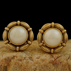 May include: A pair of round earrings featuring a central smooth white cabochon stone set within a textured antique gold-toned metal frame. The circular frame displays a repeating leaf-like pattern that surrounds the white stone, creating a classic look.