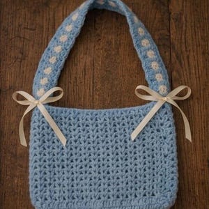 May include: A light blue crocheted bag with a single handle featuring a white polka dot pattern. The bag is accented with two cream-colored satin ribbons tied into bows on each side where the handle attaches to the main body of the bag.