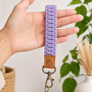 Puede incluir: Un llavero de pulsera de macramé color lavanda con un patrón de cordón tejido unido a una lengüeta de piel sintética color canela. La lengüeta tiene grabada una ilustración de hoja botánica y la letra A, unida a un mosquetón de metal bronce antiguo.