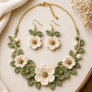 Può includere: Un set di gioielli composto da una collana e orecchini pendenti abbinati. I pezzi presentano fiori bianchi e foglie verde salvia realizzati all'uncinetto, impreziositi da piccole perline dorate al centro. La collana è fissata a una catenina dorata.