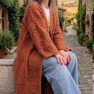 Puede incluir: Un cárdigan largo de ganchillo color óxido hecho de motivos cuadrados unidos, con mangas largas y frente abierto, usado sobre una camiseta blanca y jeans azul claro con dobladillo sin rematar, con una textura de hilo tejido a mano.