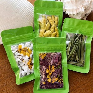 May include: Four bright green resealable pouches with clear windows, each containing different dried botanical ingredients. One bag holds yellow pods, another white and yellow petals, one purple and yellow petals, and the last green leaves.