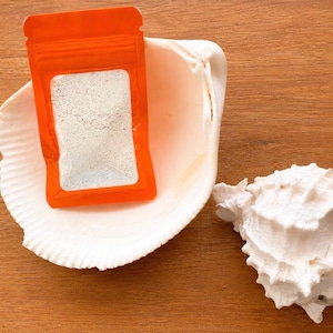 May include: A small, rectangular, bright orange resealable pouch containing a fine, off-white powder. The pouch is positioned inside a large, smooth, white seashell, next to a second, smaller, textured, white, spiky seashell.