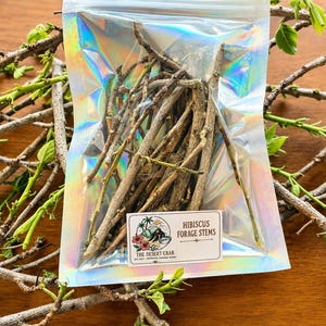 May include: A holographic plastic pouch containing brown, dried hibiscus forage stems. A white rectangular label on the front features a logo with a crab and palm trees, with the text: HIBISCUS FORAGE STEMS, THE DESERT CRAB, EST 2024.