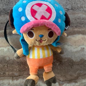 以下が含まれることがあります： 「ONE PIECE」のキャラクター、トニートニー・チョッパーのぬいぐるみリュック。キャラクターは白の水玉模様とピンクの十字架がついた青い帽子をかぶり、ベージュの顔、黄色と白のストライプのシャツ、オレンジ色のズボン、茶色のひづめが特徴です。