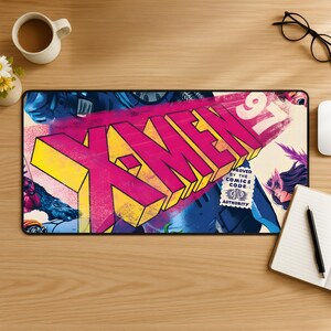 Könnte beinhalten: Eine rechteckige Schreibtischunterlage mit einer lebendigen X-Men '97-Grafik und kräftigen gelben und magentafarbenen 3D-Buchstaben. Das Design enthält Comic-Figuren, ein Comics Code Authority-Siegel und einen schwarzen Rand.