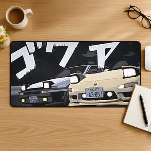 Könnte beinhalten: Eine rechteckige Schreibtischunterlage mit einer Anime-Illustration von zwei Sportwagen mit Klappscheinwerfern. Das linke Auto ist weiß und schwarz, das rechte cremefarben. Große weiße japanische Katakana-Schriftzeichen sind über den Autos zu sehen.