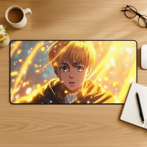 Op de afbeelding: Een rechthoekige muismat met een anime-personage met blond haar en blauwe ogen, tegen een achtergrond van gouden licht. Het personage draagt een bruine en groene jas met capuchon. De muismat ligt op een houten bureau met een kopje koffie en andere kantoorbenodigdheden.