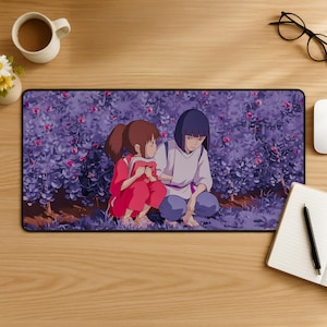 Könnte beinhalten: Ein rechteckiges Mauspad mit einer Anime-Szene mit zwei Mädchen in einer lila Blumeneinstellung. Ein Mädchen trägt ein rotes Outfit, das andere ein weißes und blaues Outfit. Das Mauspad liegt auf einem Holztisch mit einer Kaffeetasse und Büromaterial.