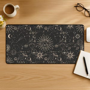 Könnte beinhalten: Eine schwarze, rechteckige Schreibtischunterlage mit einem himmlischen Design aus Sonnen, Monden, Sternen und Sternbildern in Creme. Die Unterlage ist von Büromaterialien umgeben, darunter eine Kaffeetasse, eine Brille und ein Notizbuch.