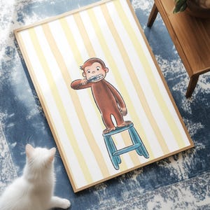 Puede incluir: Una lámina de arte enmarcada que muestra a un mono marrón de pie sobre un taburete azul mientras se cepilla los dientes. El fondo consiste en rayas verticales de color amarillo pálido y blanco. La ilustración está centrada en un marco de madera clara.