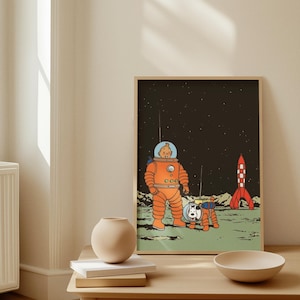 May include: A framed illustration featuring a cartoon astronaut in an orange spacesuit with a dog in a matching suit on a moon-like surface. A red and white rocket is in the background against a starry sky. The artwork is displayed on a light-colored wall.