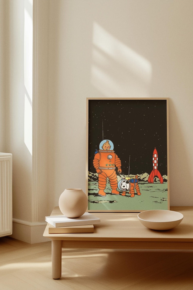 May include: A framed illustration featuring a cartoon astronaut in an orange spacesuit with a dog in a matching suit on a moon-like surface. A red and white rocket is in the background against a starry sky. The artwork is displayed on a light-colored wall.