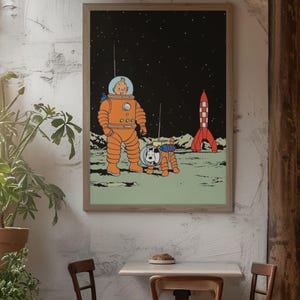 May include: Framed print featuring a cartoon illustration of a character in an orange spacesuit with a dog in a spacesuit on a moon-like surface. A red and white rocket is in the background against a starry sky. The print is displayed on a white wall.