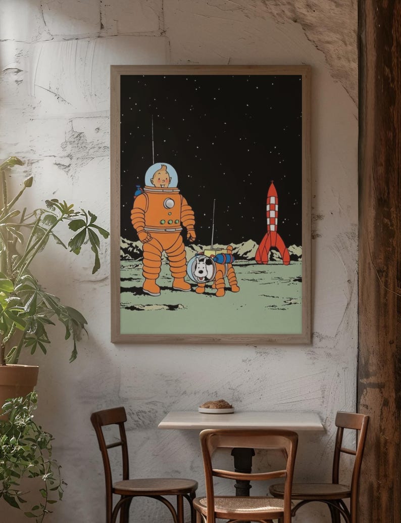 May include: Framed print featuring a cartoon illustration of a character in an orange spacesuit with a dog in a spacesuit on a moon-like surface. A red and white rocket is in the background against a starry sky. The print is displayed on a white wall.