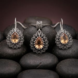 Op de afbeelding: Een sieradenset bestaande uit een hanger en twee oorbellen, elk voorzien van een peervormige, licht amberkleurige edelsteen in een zilverkleurige metalen zetting met ingewikkeld filigraan krulwerk en een textuur op de hanger.