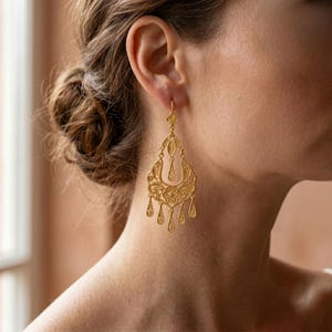 Könnte beinhalten: Ein goldfarbener Hängeohrring mit einem komplizierten Filigranmuster, einem zentralen Blumenmotiv und mehreren kleinen tropfenförmigen Anhängern an der Unterkante. Der Ohrring besteht aus einer feinen, durchbrochenen Metallstruktur.