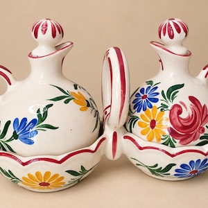 May include: A ceramic oil and vinegar cruet set featuring two round bottles with handles and matching stoppers, resting in a connected double-bowl base. The white glazed surface is hand-painted with vibrant red, yellow, and blue floral motifs.