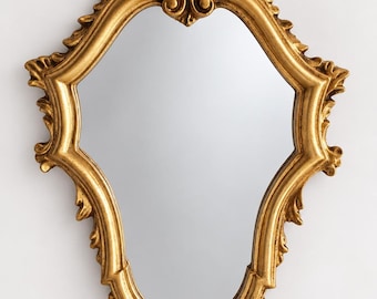 Peut inclure: Un miroir mural avec un cadre doré présentant une forme ornée rappelant un bouclier. Le cadre est décoré de volutes complexes et de sculptures en forme de feuilles en haut et en bas, entourant une surface en verre réfléchissante et lisse.