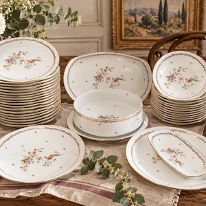 Peut inclure: Un grand service de table en porcelaine blanche orné d'un motif floral et d'oiseaux délicat avec des bords dorés. La collection comprend des assiettes empilées, un bol de service profond, un plat rectangulaire et des plats ovales.