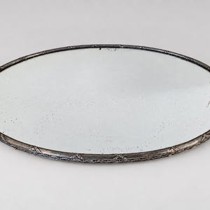 Op de afbeelding: Een ovale spiegel met een donker metalen frame met een gestructureerd, herhalend patroon langs de buitenrand. Het reflecterende oppervlak is helder en glad, omlijst door de gedetailleerde, donkere metalen rand die het hele stuk omsluit.
