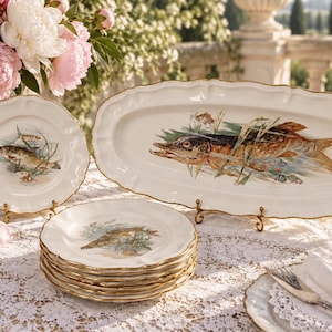 Op de afbeelding: Een set wit keramieken servies met een visillustratie, groen riet en goudkleurige, geschulpte randen. De collectie bestaat uit een grote ovale serveerschaal, een enkel rechtopstaand bord en een stapel van vijf bijpassende dinerborden.